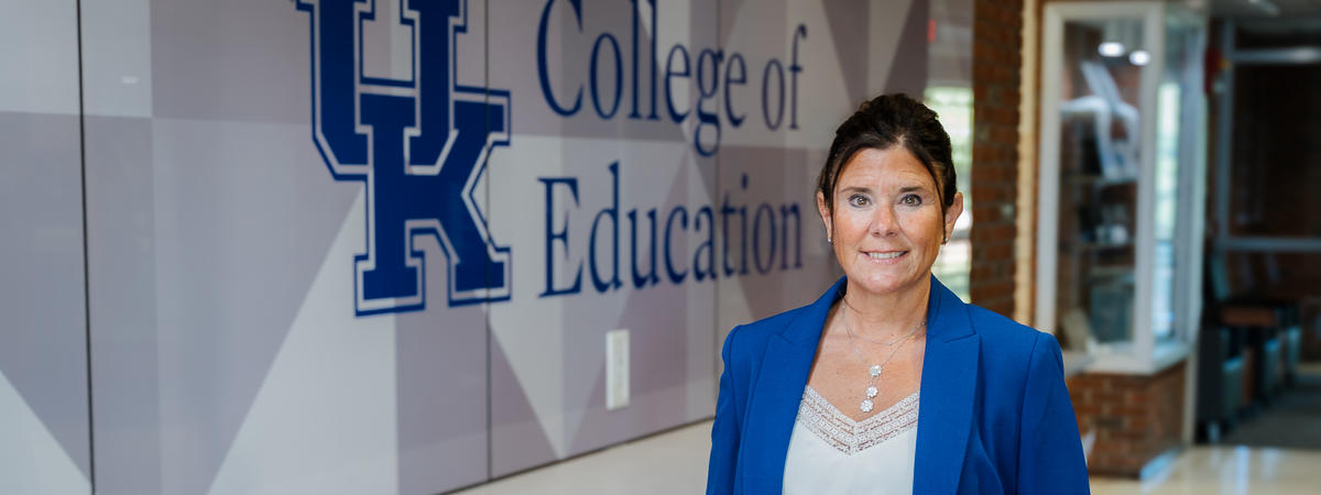 A person in a blue suit stands in a hallway beside a wall displaying the University of Kentucky logo and the words “College of Education.” A counter with stools lines the wall, and the hallway extends into the background.