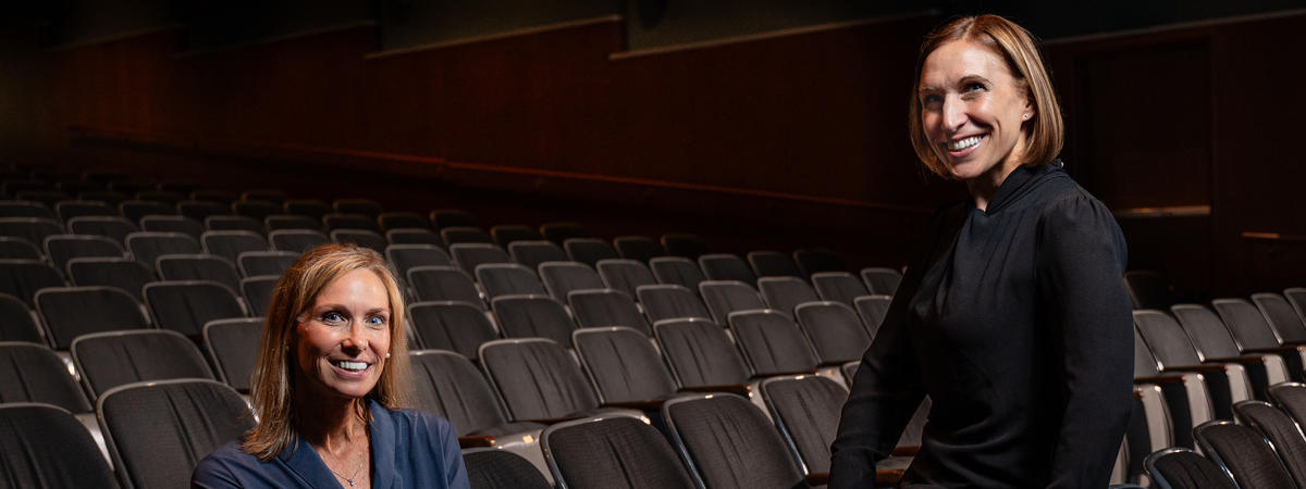 Two people pose among rows of seats in a dimly lit auditorium, with one seated and the other leaning against a nearby chair. Wall lights illuminate the theater in the background.
