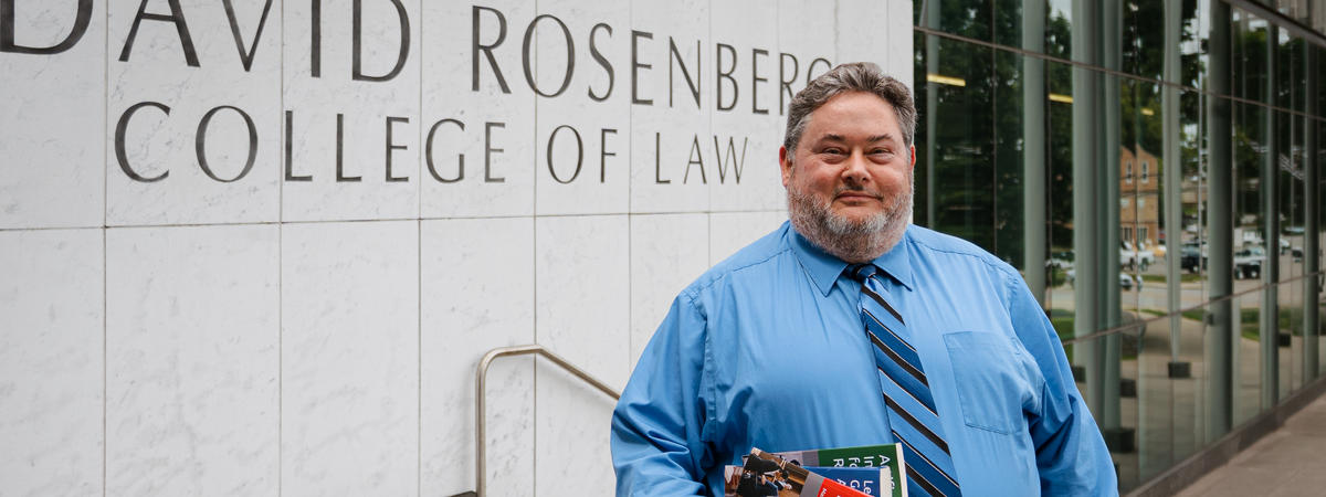 A person stands outside the Rosenberg College of Law building, holding several books.