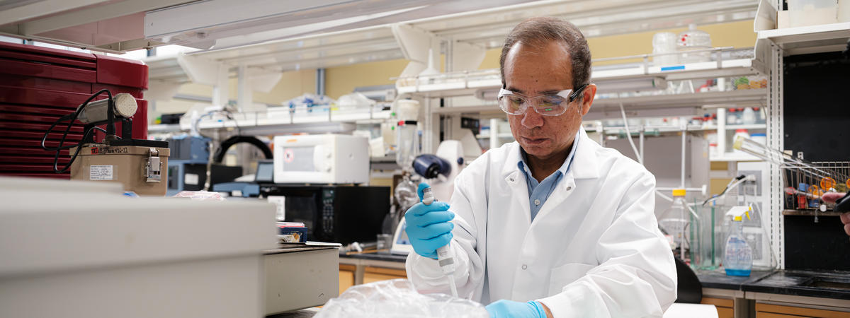 A researcher wearing a white lab coat, safety glasses and blue gloves uses a pipette while working at a laboratory bench surrounded by scientific instruments, bottles and testing equipment in a University of Kentucky research lab.