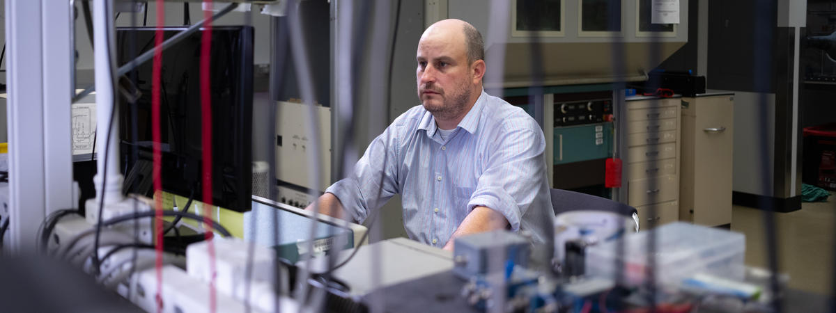 A researcher sits at a computer workstation in a laboratory, viewed through an array of cables, instruments and scientific equipment. 