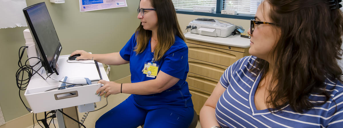 A health care professional in blue scrubs uses a computer beside a seated pregnant patient in an exam room.