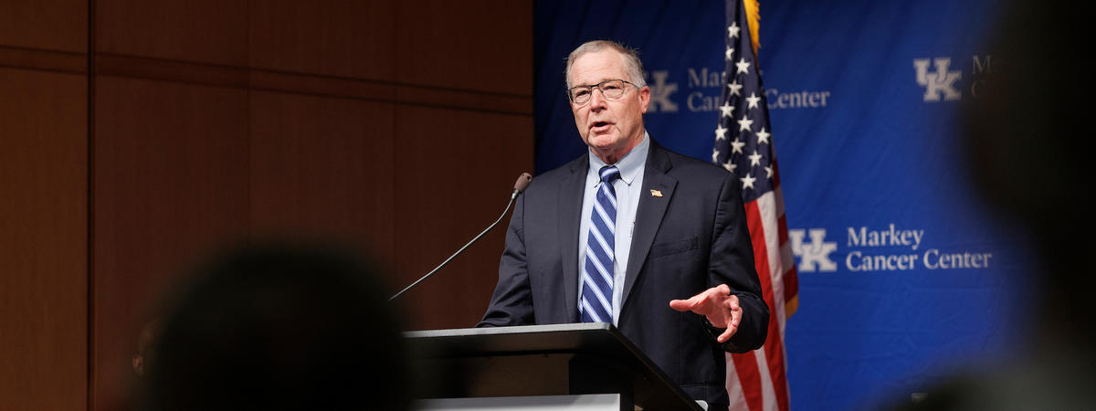 A person wearing glasses, a dark suit and striped blue tie speaks at a podium during a formal event. An American flag and a blue backdrop displaying the UK Markey Cancer Center logo are behind the speaker.