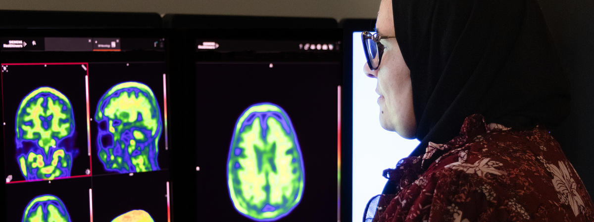 A woman wearing glasses and a headscarf studies colorful brain scan images displayed on two computer monitors in a medical research setting.