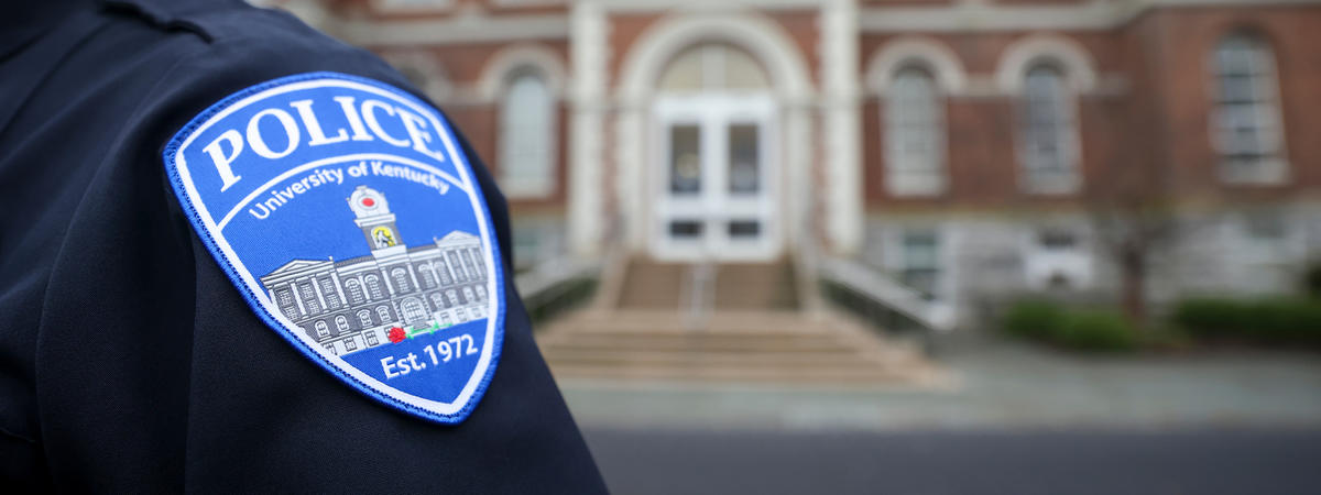 Police uniform with a University of Kentucky Police Department shoulder patch in the foreground, with a campus building blurred in the background.  Provide your feedback on BizChat