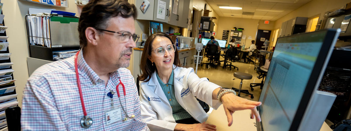 Two health care providers review information on a computer workstation in a hospital clinic. One points to data on the screen while discussing results, illustrating collaboration in patient care and clinical decision-making.