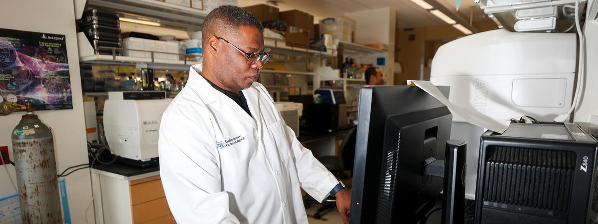 Person wearing a white lab coat, works at a computer beside research equipment in a laboratory.
