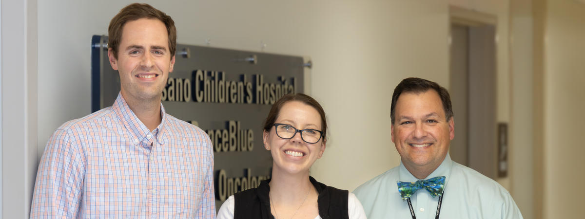 Three healthcare team members stand together in a hospital hallway in front of a sign for a pediatric oncology clinic. One person wears a plaid button-down shirt, another wears a black hospital vest with an ID badge, and the third wears a light blue shirt