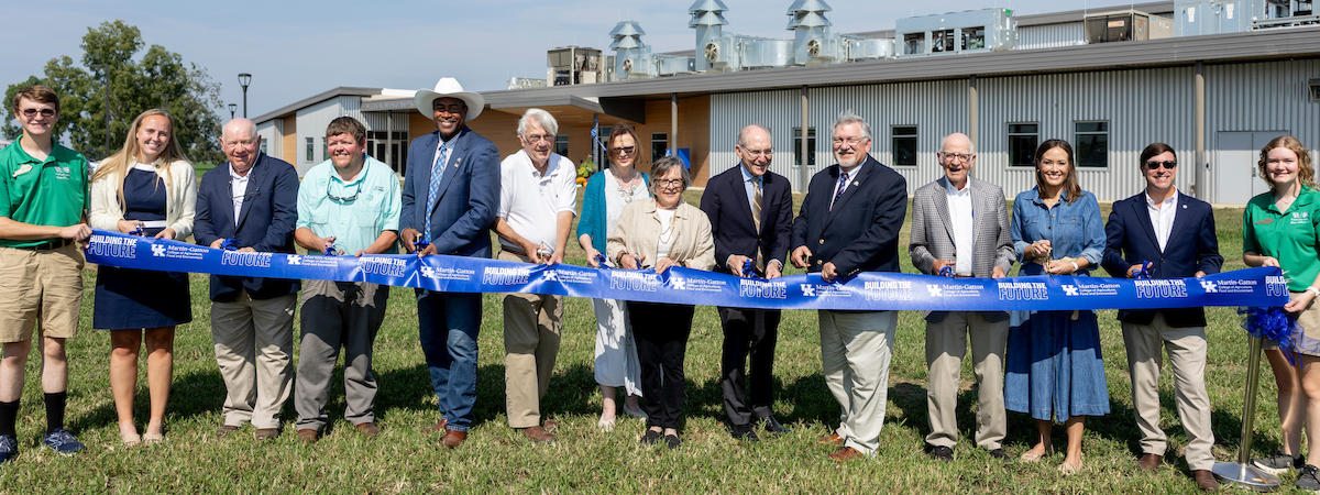 A group of people standing outdoors in front of a large modern industrial or educational facility, holding a blue ceremonial ribbon and scissors during a ribbon-cutting event. The building features metal siding and rooftop ventilation equipment.
