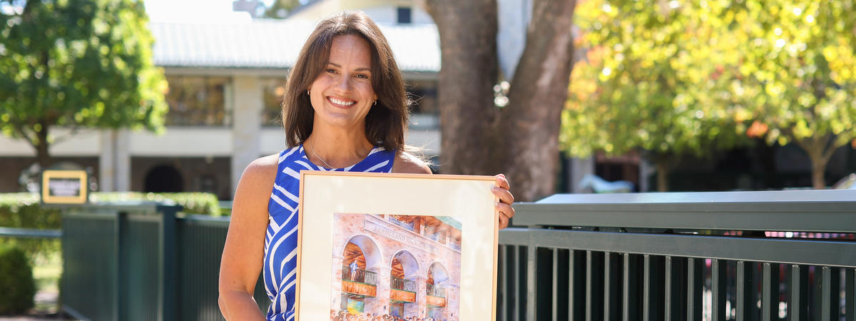 A smiling person in a blue-and-white sleeveless top stands outdoors holding a framed painting of a horse and spectators.