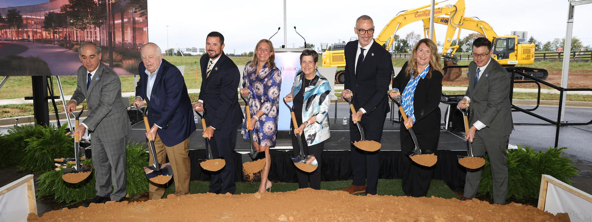 A group of people wearing hard hats pose behind a ceremonial dirt pile with shovels at a groundbreaking event under a large white tent. A banner along the front displays the UK HealthCare logo and the names Perkins&Will and Walsh.