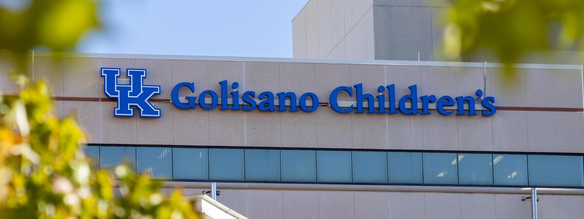 Exterior of the Golisano Children’s building featuring the blue UK logo and “Golisano Children’s” sign, framed by green tree leaves against a blue sky.