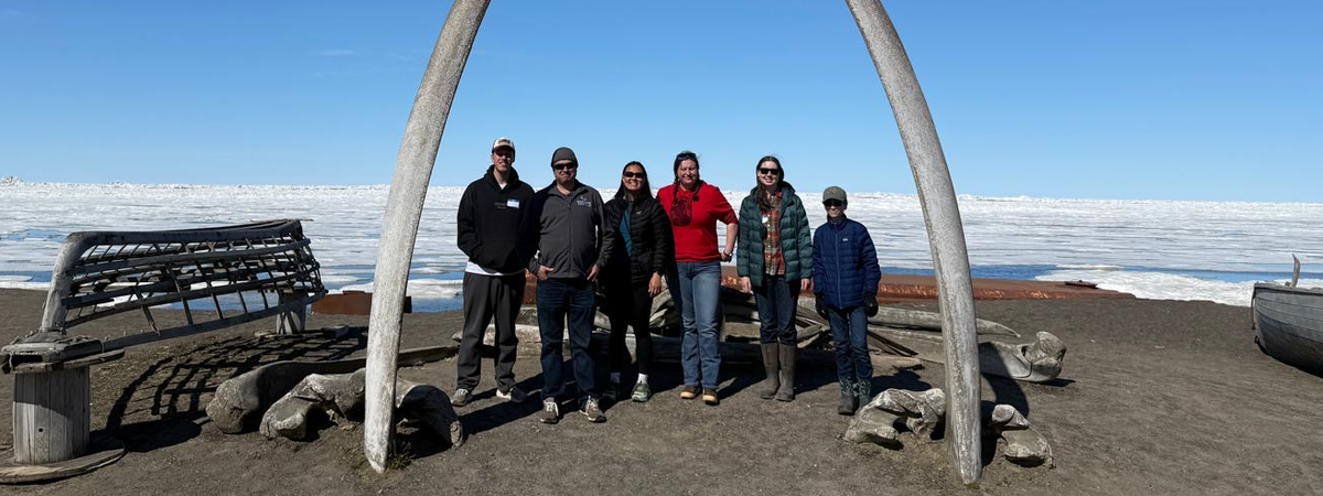 Six people stand beneath a large arch formed by two whale bones on a dark shoreline, with wooden boats and a vast expanse of sea ice beneath a clear blue sky in the background.