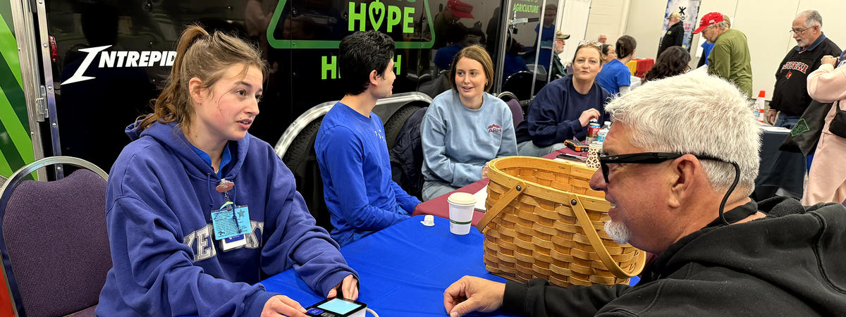 People sit around a blue-covered table at a community event, engaging in an activity with small red-and-white items and a basket on the table.