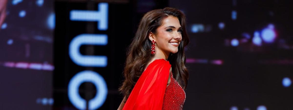 Person in a sparkling red gown with a flowing cape walks across a stage, with a large "Kentucky" sign and dark, illuminated backdrop in the background. 
