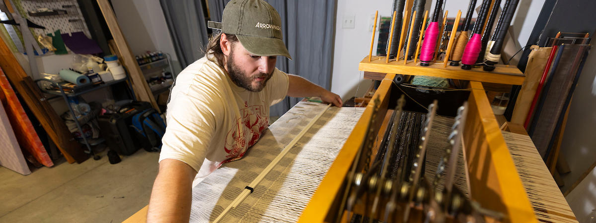 White male in a white t-shirt wearing a ball cap, seated at a loom. 