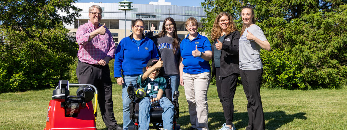 Group photo outdoors featuring a child seated in a motorized wheelchair surrounded by six adults. A red adaptive football launcher is parked beside the group, with trees and a university building visible in the background.