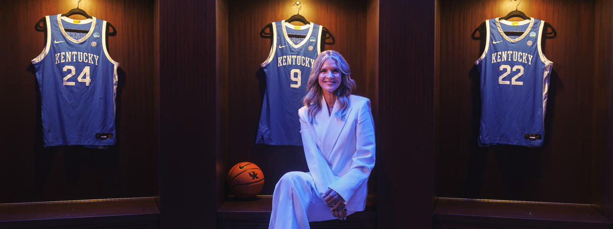 A person dressed in a light-colored suit sits in the center of a display featuring framed University of Kentucky basketball jerseys. Three blue jerseys, including numbers 24, 3 and 22, are mounted in illuminated wooden display cases behind the person.