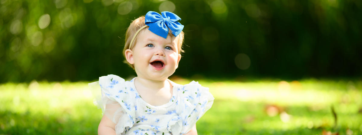 A young child sits on green grass outdoors, wearing a white floral outfit and a large blue bow headband. The background is softly blurred with trees and sunlight filtering through the greenery.
