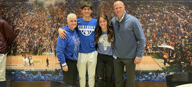 Four people pose together in front of a large mural depicting a University of Kentucky basketball championship celebration. The group stands with their arms around one another, wearing Kentucky-themed clothing, confetti and a packed arena crowd in backgro
