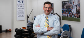 Person stands with arms crossed in a biomechanics lab containing dumbbells, exercise equipment and wall displays, including a sign reading, “We cannot walk alone.”