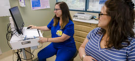 A health care professional in blue scrubs uses a computer beside a seated pregnant patient in an exam room.