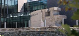 Exterior view of a modern campus building with large glass windows and stone walls, featuring a tiered stone landscape with green shrubs in the foreground and architectural details of the student center visible behind.