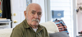 A man sits on a couch in a living room, wearing an olive-green shirt with a phone in the chest pocket.