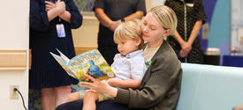 Adult sits on a bench reading a picture book to a young child on their lap, while several people stand nearby in a hospital-like setting.