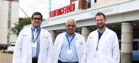 Three physicians wearing white lab coats stand outside the emergency department entrance at a hospital. The group poses together in front of the emergency care facility, highlighting a clinical and academic health care setting.