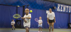 A child swings a tennis racquet to hit a bright yellow tennis ball on an indoor court at the University of Kentucky. Three other young tennis players and an instructor stand in the background holding racquets. Blue court curtains and large “KENTUCKY” lett