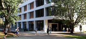 Students walk along a shaded campus pathway outside a modern academic building surrounded by large trees.