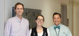 Three healthcare team members stand together in a hospital hallway in front of a sign for a pediatric oncology clinic. One person wears a plaid button-down shirt, another wears a black hospital vest with an ID badge, and the third wears a light blue shirt