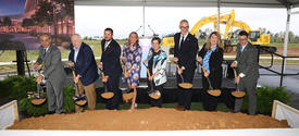 A group of people wearing hard hats pose behind a ceremonial dirt pile with shovels at a groundbreaking event under a large white tent. A banner along the front displays the UK HealthCare logo and the names Perkins&Will and Walsh.