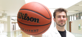 Man wearing a white lab coat holds a Wilson basketball prominently in front of them inside a bright, modern building.