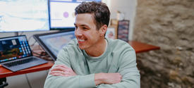 Person in a light green sweatshirt sits on a wooden chair with arms crossed in front of a desk with a laptop and two large monitors; stone wall in the background.