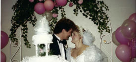 Photo of a young white couple at their wedding. A multi-tiered cake is in the foreground; the couple stand behind it, facing each other with foreheads touching and big smiles on their faces. They're framed by a canopy of foliage and pink balloons. 