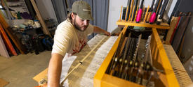 White male in a white t-shirt wearing a ball cap, seated at a loom. 