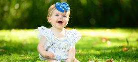 A young child sits on green grass outdoors, wearing a white floral outfit and a large blue bow headband. The background is softly blurred with trees and sunlight filtering through the greenery.