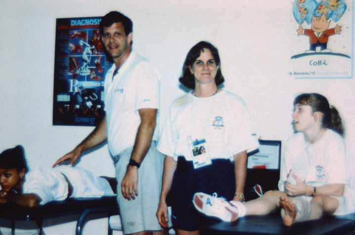 Dr. Ireland stands in the middle of the screen to her right a female gymnast sits on a table, to her left a male doctor stands by a female gymnast laying on a table.