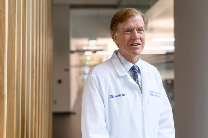 A physician in a white UK HealthCare lab coat smiles while standing in a modern medical facility hallway.