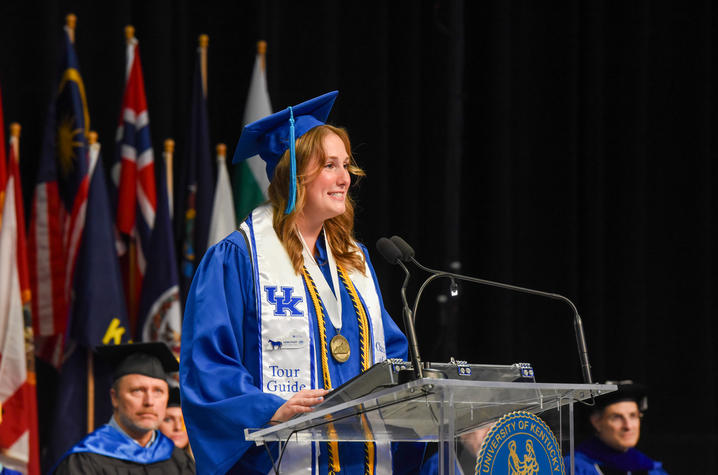 photo of May 2026 UK commencement A graduate stands at a podium in cap and gown with honor cords and medal, speaking during a commencement ceremony with flags in the background.
