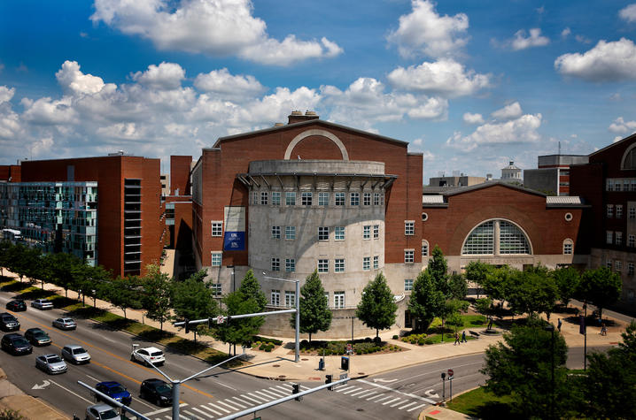This is an exterior photo of the Charles T. Wethington Jr. Building, a large, rounded pale stone and red brick facade at the corner of two streets, with trees in front and a pale blue sky dotted with clouds. 