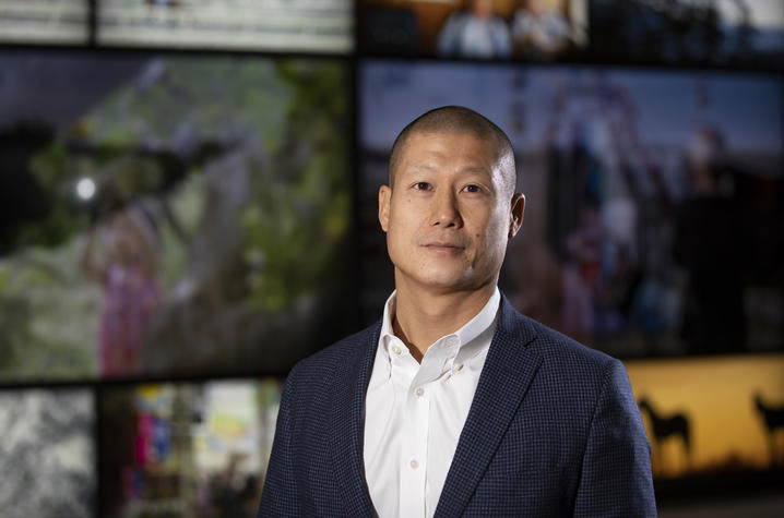 James Liau, M.D. Portrait of a person wearing a white dress shirt and blue checkered blazer, standing in front of a wall of blurred display screens.