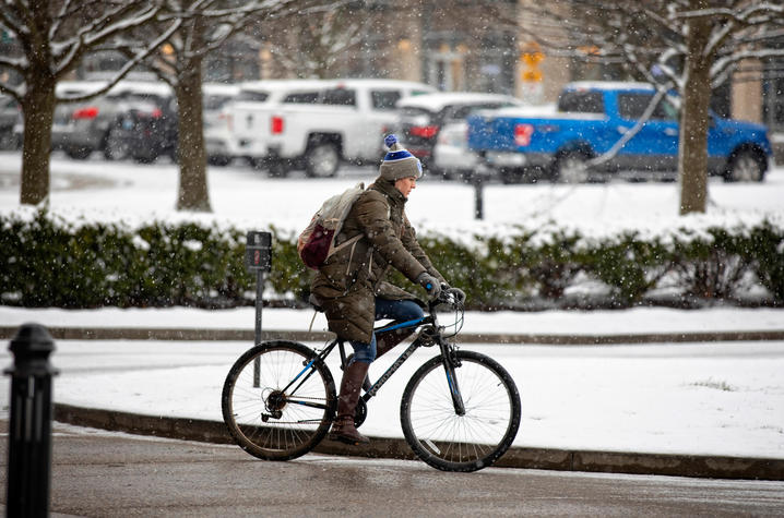 A person rides a bicycle along a snow-covered street during snowfall.