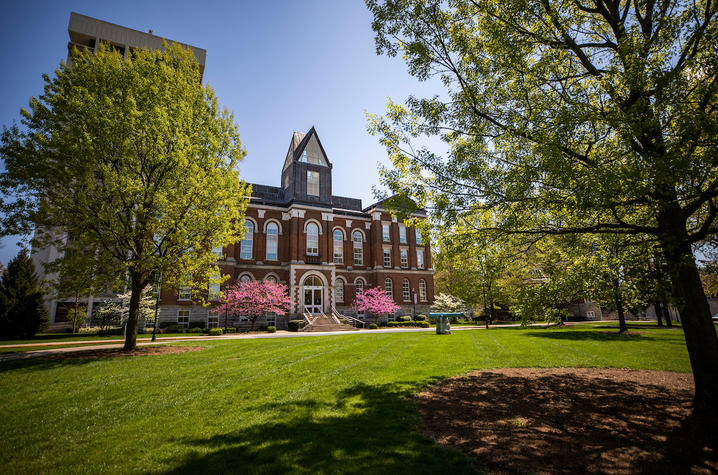 The front of Main Building at the University of Kentucky during the springtime, with trees and grass in the foreground and a clear blue sky in the background.