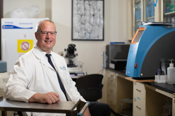 A man wearing a white lab coat sits in a laboratory beside scientific equipment and smiles toward the camera.