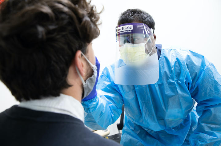 A member of the study team, wearing a protective gown, face shield, and face mask, takes a nose swab of a young man whose back is to the viewer. 