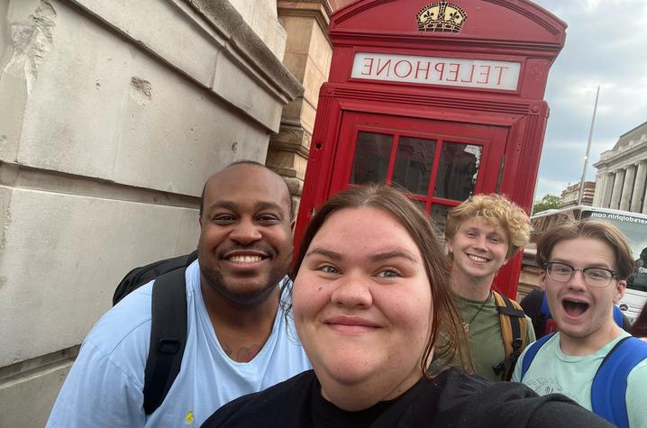 Four people take a close-up selfie on a city street, with a bright red British-style phone booth behind them and stone buildings lining the sidewalk.