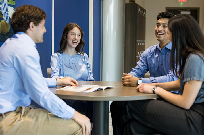 Four people sit around a small round table in an office setting, engaged in a discussion with documents on the table, suggesting a meeting or collaborative conversation.